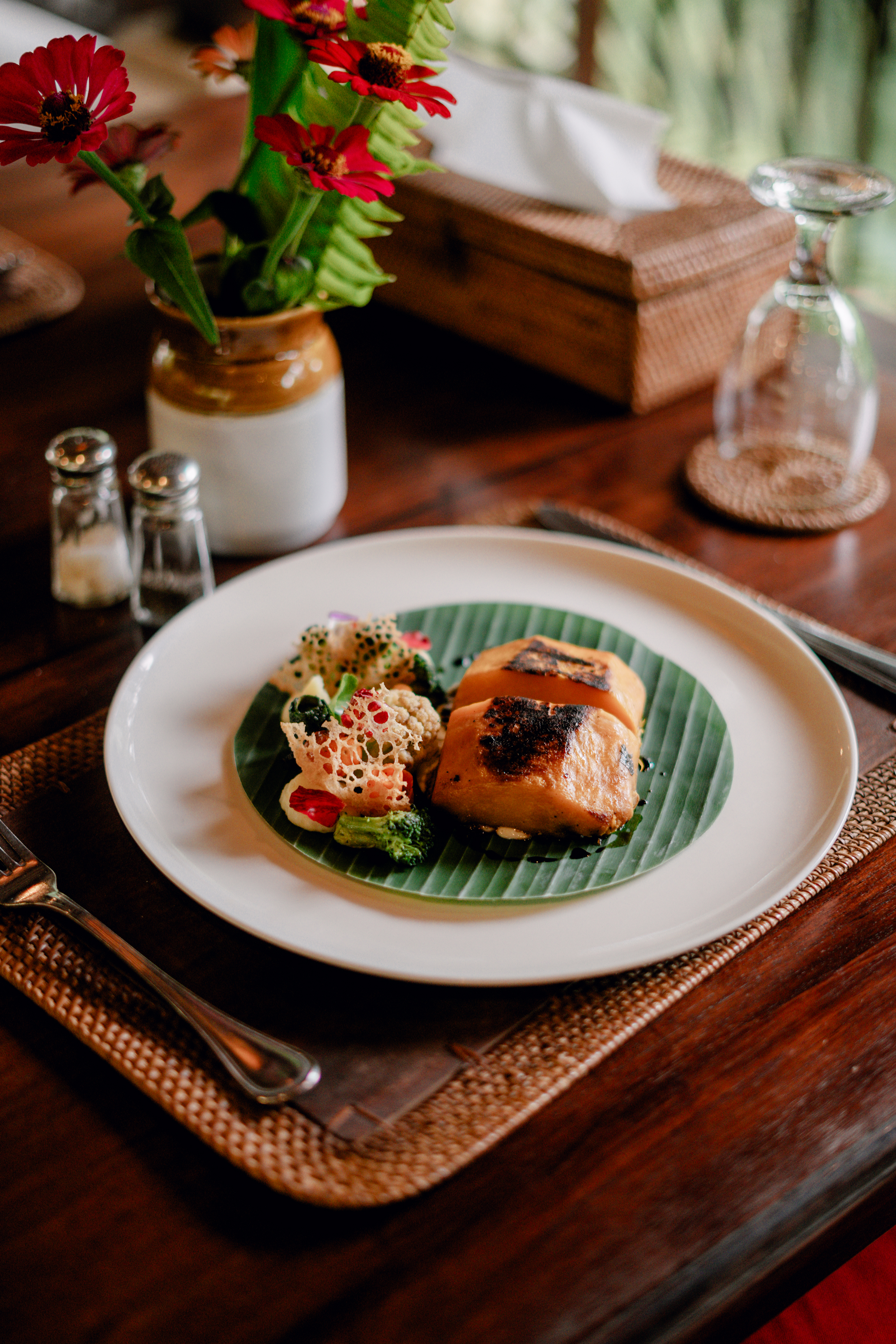 Plant-based dish served on a banana leaf at a teak dining table with fresh flowers